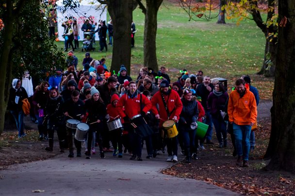 Photo of a group of people walking in a park. Some of the group are playing drums as they walk.