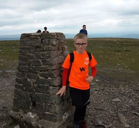 Photo of a young boy wearing an orange Barnsley Hospice t-shirt. He is standing at the summit of Ingleborough which is a stone built cairn marker