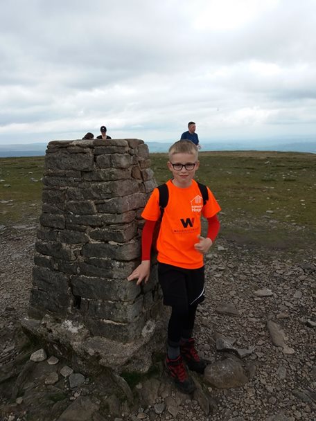 Photo of a young boy wearing an orange Barnsley Hospice t-shirt. He is standing at the summit of Ingleborough which is a stone built cairn marker