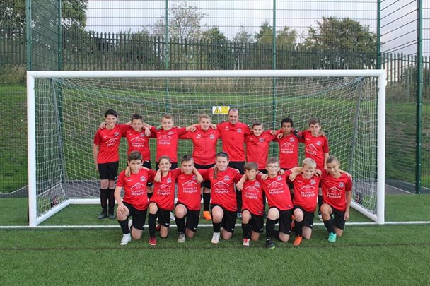 Photo of a children's football team wearing red shirts. The group are standing in two rows in a football goal post.