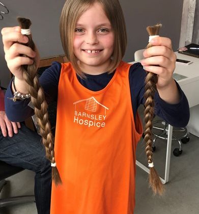 Photo of a young girl wearing an orange Barnsley Hospice vest. The girl is holding her pigtails in her hands which she has cut off to raise money for the Hospice.