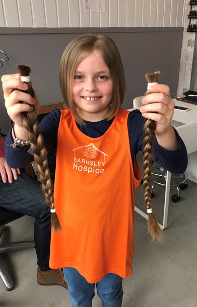 Photo of a young girl wearing an orange Barnsley Hospice vest. The girl is holding her pigtails in her hands which she has cut off to raise money for the Hospice.