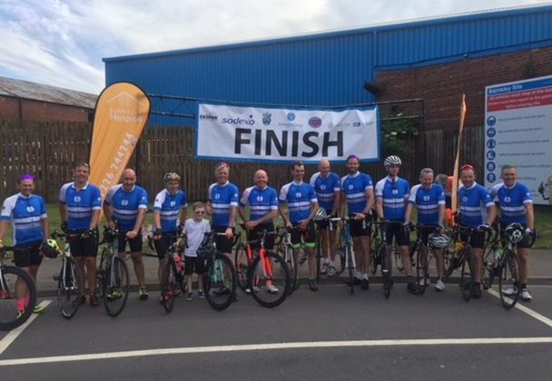 Photo of a line of cyclists and their bikes. All the cyclists are wearing blue shirts and there is a large finish banner behind them.