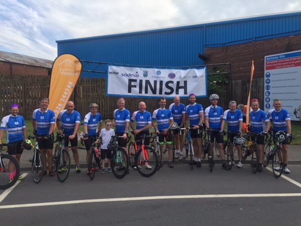 Photo of a line of cyclists and their bikes. All the cyclists are wearing blue shirts and there is a large finish banner behind them.