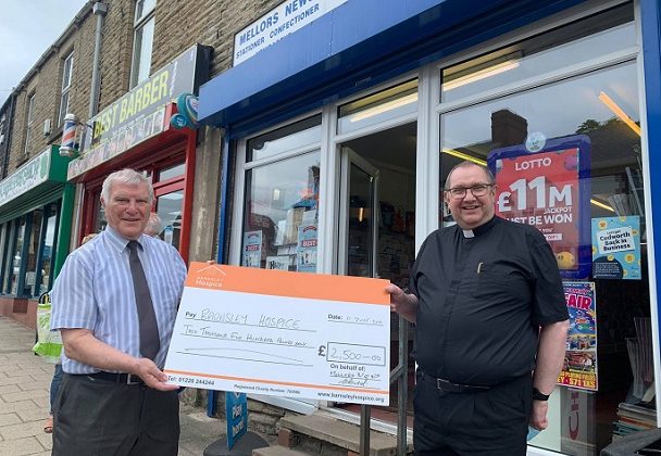 Photo of newsagent Gordon Bird and Father David holding a donation cheque for Barnsley Hospice. They are standing outside Gordon's newsagent.