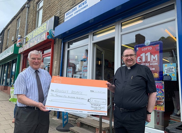 Photo of newsagent Gordon Bird and Father David holding a donation cheque for Barnsley Hospice. They are standing outside Gordon's newsagent.