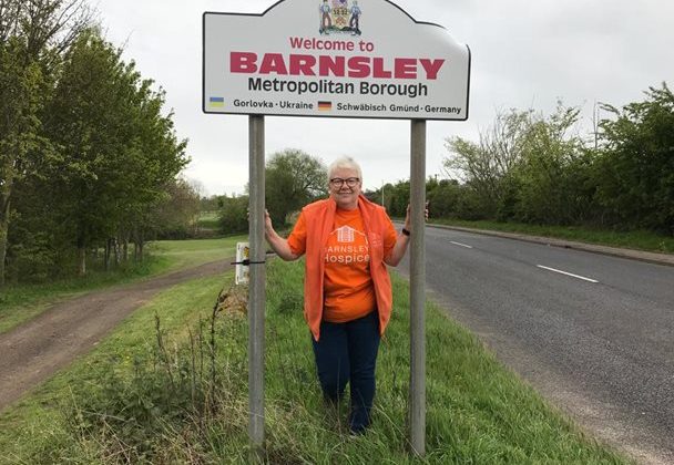 Photo of a lady wearing an orange coloured Barnsley Hospice t-shirt standing under the Welcome to Barnsley road sign