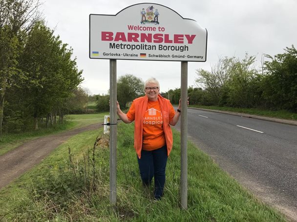 Photo of a lady wearing an orange coloured Barnsley Hospice t-shirt standing under the Welcome to Barnsley road sign