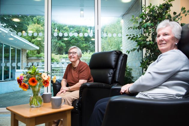 Photo of two ladies seated in the Barnsley Hospice orangery. The ladies are smiling and seated in comfortable armchairs with a view of the garden