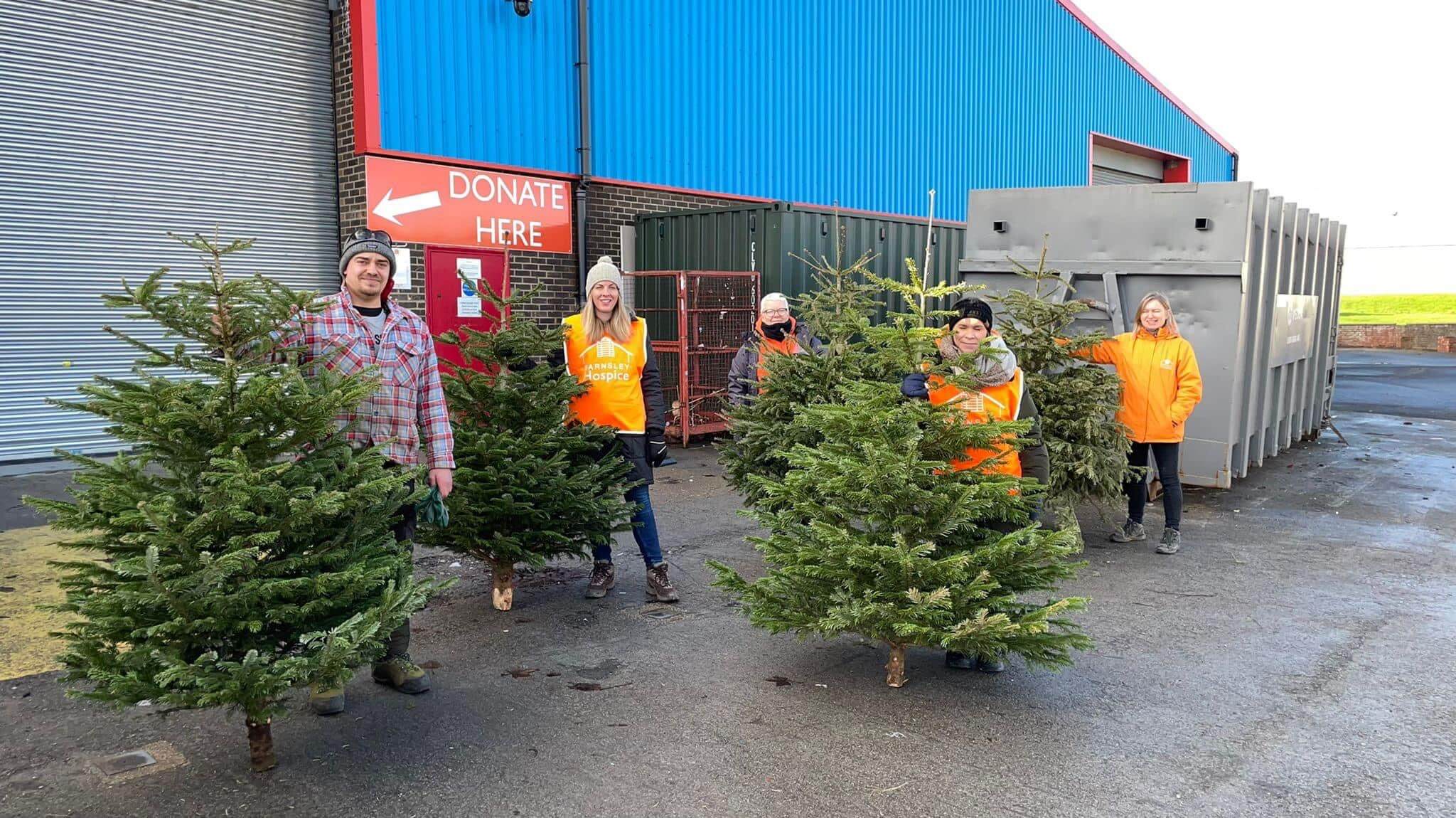 Photo of five members of the Barnsley Hospice team standing in front of the donations warehouse. They are holding Christmas trees as they assist with the Treecycle fund raising event
