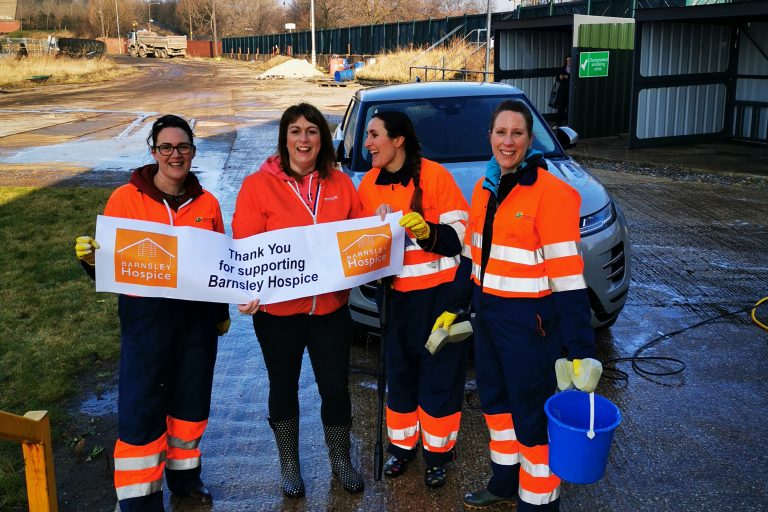 Photo of four ladies wearing waterproof clothing, holding a bucket and a Barnsley Hospice banner after taking part in a car wash fundraising event.