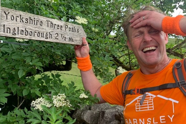 Hospice fundraiser looking tired, but smiling. He is holding onto a Yorkshire Three Peaks sign that says 