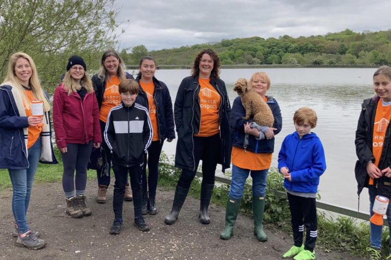 A group of fundraisers wearing orange hospice t-shirts. They are stood. together smiling in front of a reservoir. One lady is carrying a small brown dog.
