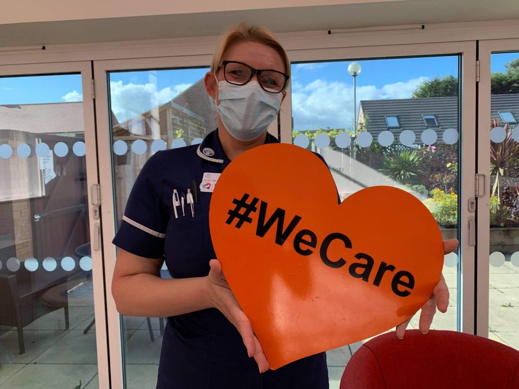 A hospice nurse smiling holding a orange heart that reads #WeCare. She is wearing navy nurses uniform and a facemask,