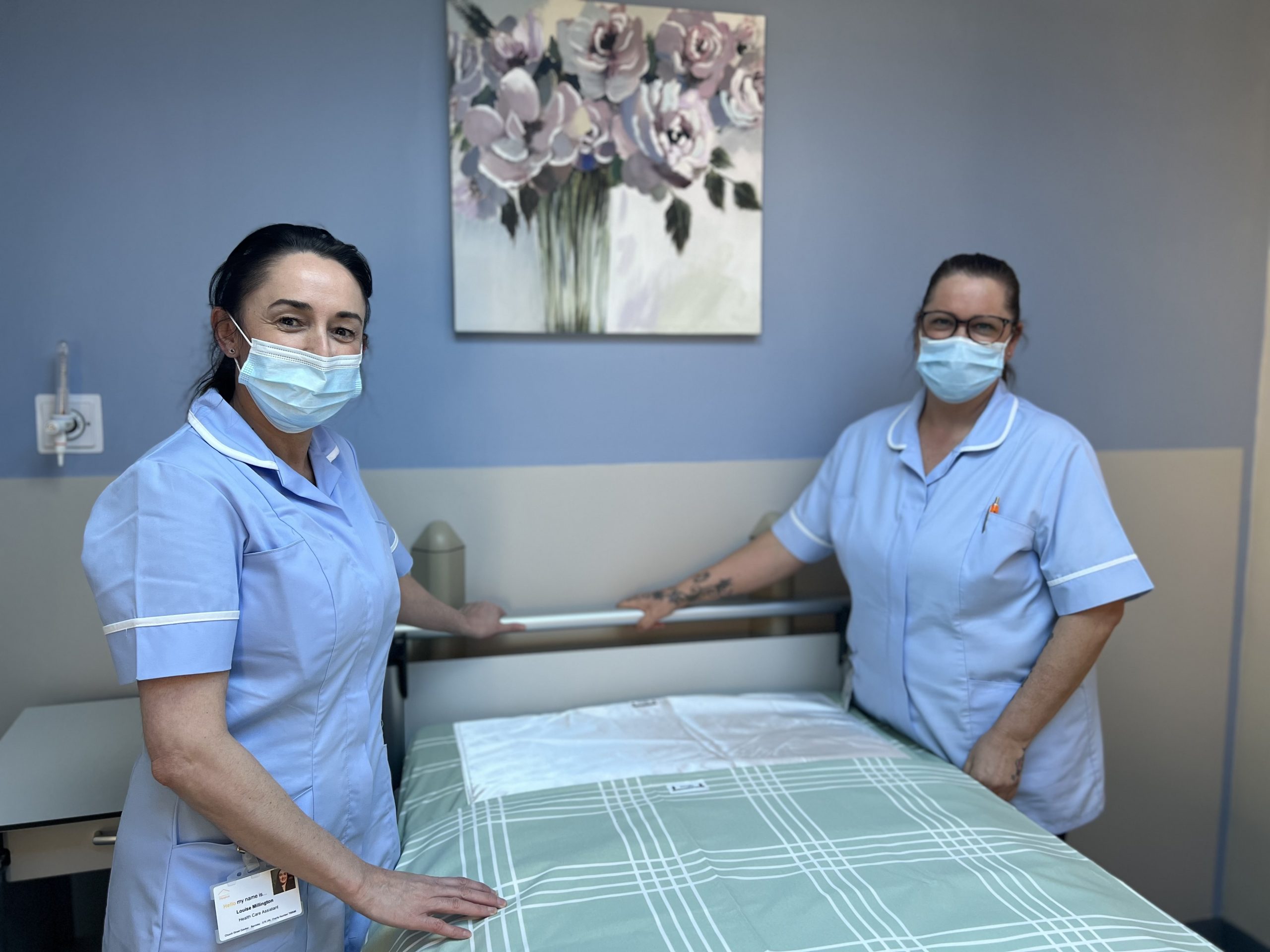 Two healthcare assistants making a bed on the inpatient unit. The bedding is light green and there is a picture of pink flowers on the wall behind. The two members of staff are smiling and are dressed in light blue uniform. They are wearing face masks.