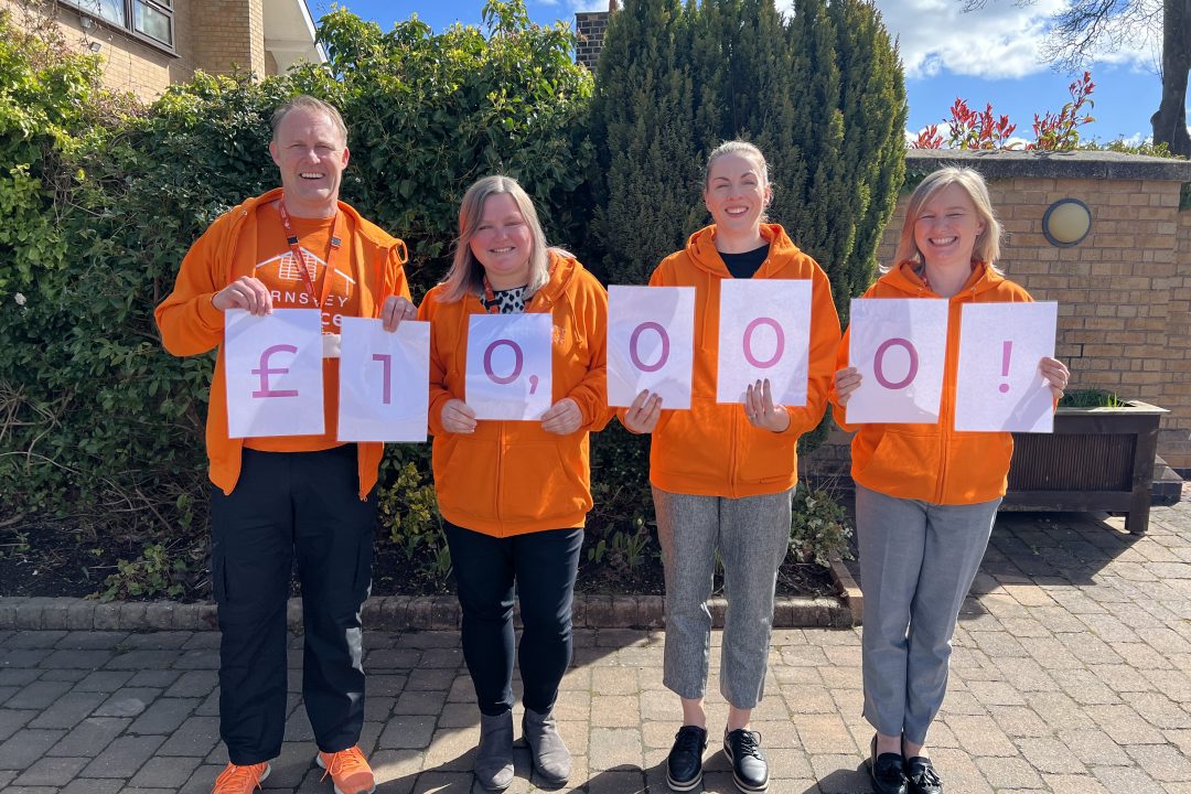 Four hospice fundraisers holding numbers that spell out £10,000. The fundraising team are wearing orange hospice jumpers and are posing outside the hospice.