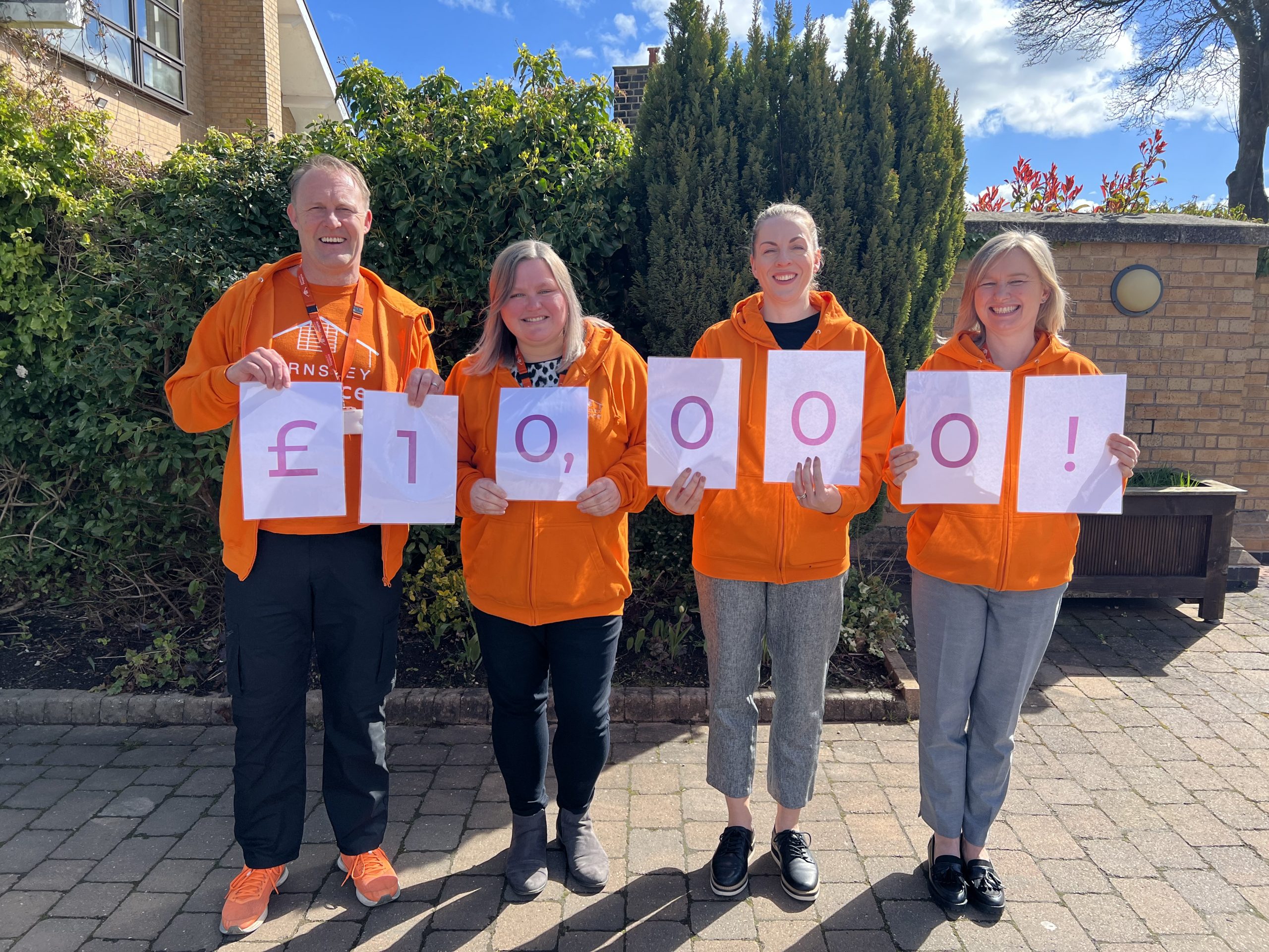 Four hospice fundraisers holding numbers that spell out £10,000. The fundraising team are wearing orange hospice jumpers and are posing outside the hospice.