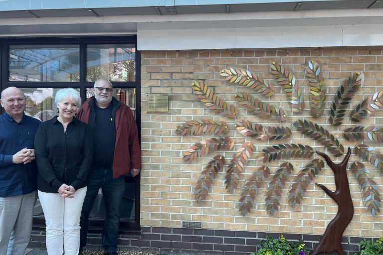 Supporters of the hospice smiling outside the Legacy Tree. The Legacy Tree is on the front of the hospice. It is an artwork shaped like a tree with a wooden sculpture as the trunk with individual metal leaves. The leaves are gold, silver and bronze.