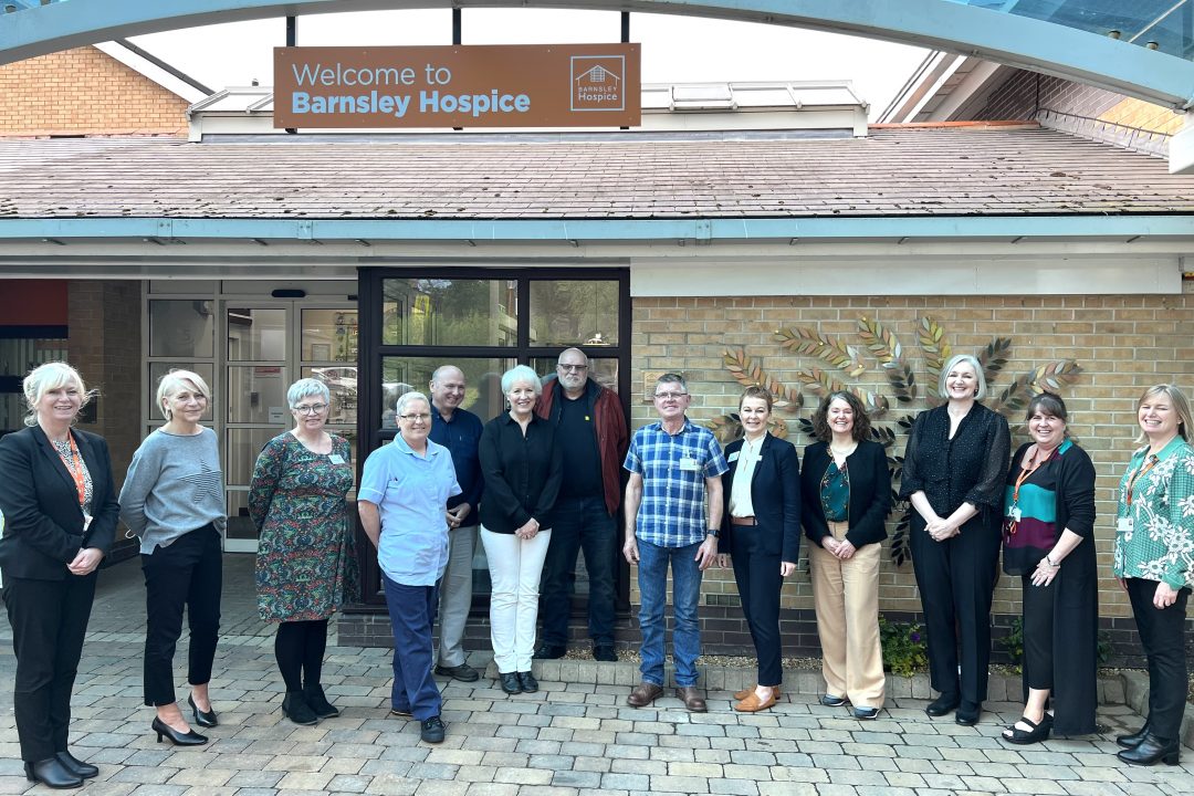 A team photo of staff and supporters smiling outside the front of the hospice. You can see The Legacy tree in the background.