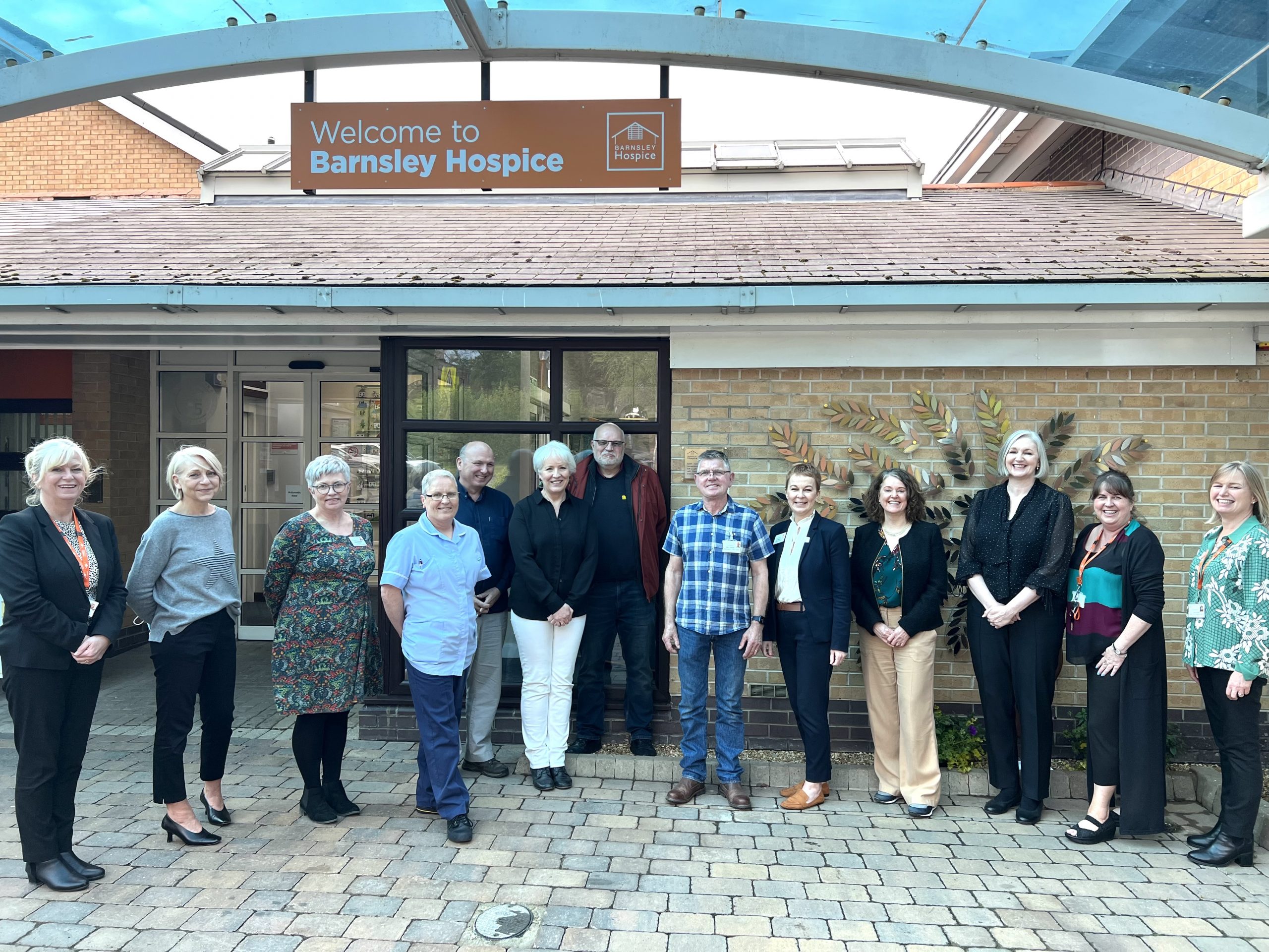 A team photo of staff and supporters smiling outside the front of the hospice. You can see The Legacy tree in the background.