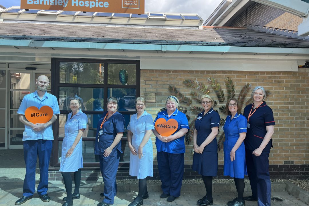 Eight members of the hospice nursing staff smiling outside the front of the hospice. The team are in their nursing uniform and are stood in line. Two members of staff are holding orange hearts that say #WeCare.