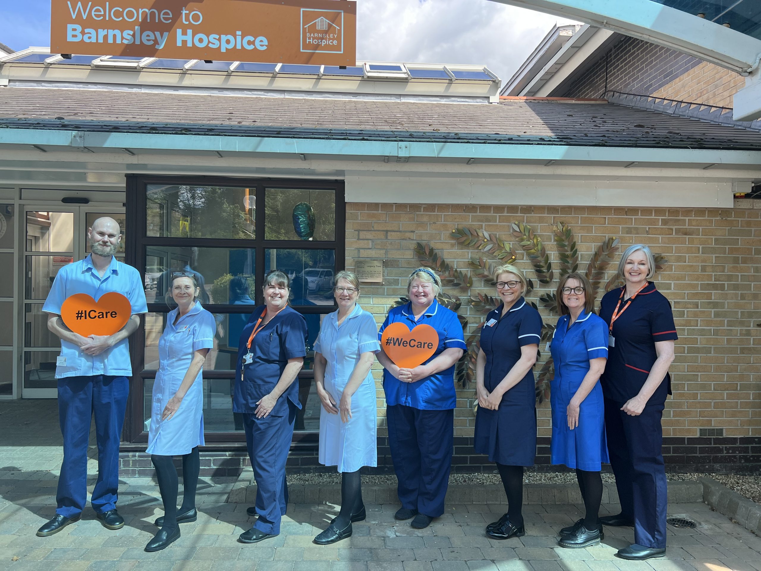 Eight members of the hospice nursing staff smiling outside the front of the hospice. The team are in their nursing uniform and are stood in line. Two members of staff are holding orange hearts that say #WeCare.