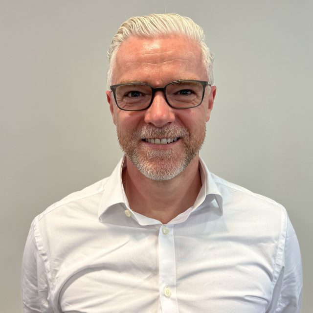 Headshot of Director of Human Resources and Organisational Development, Stephen Hall. Stephen has white hair and is smiling at the camera. He wears black-framed glasses and is wearing a white button-up shirt.
