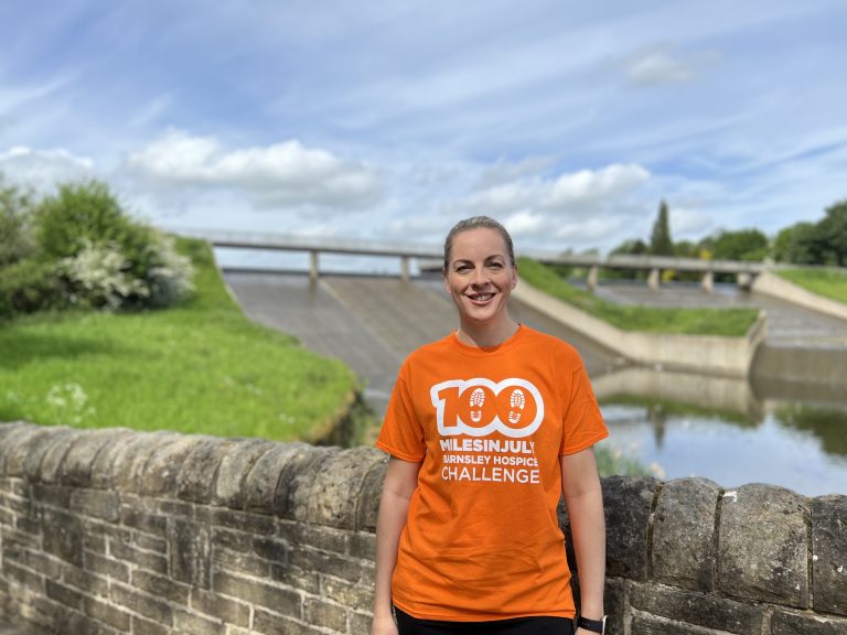 Lady wearing orange t-shirt saying 100 Miles in July t in front of a reservoir.