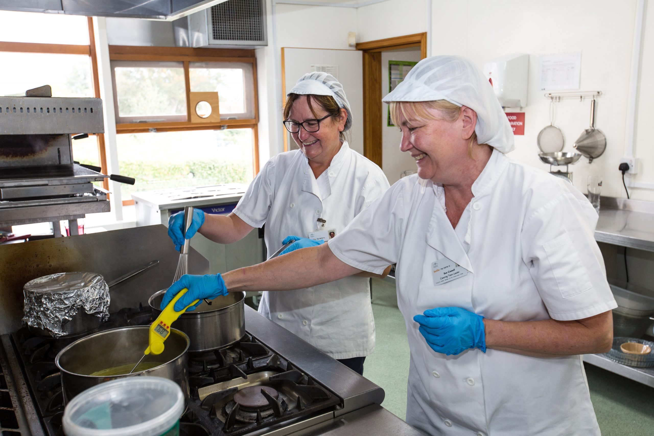 Photo of two ladies from the catering team in the kitchen at Barnsley Hospice. They are dressed in white catering outfits and preparing food on a hob.