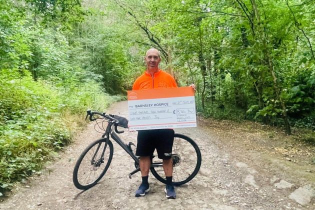 Photo of fundraiser Gavin Coles holding a donation cheque. Gavin is pictured with his bicycle standing on a woodland path.