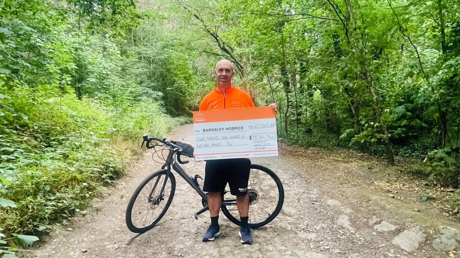 Photo of fundraiser Gavin Coles holding a donation cheque. Gavin is pictured with his bicycle standing on a woodland path.