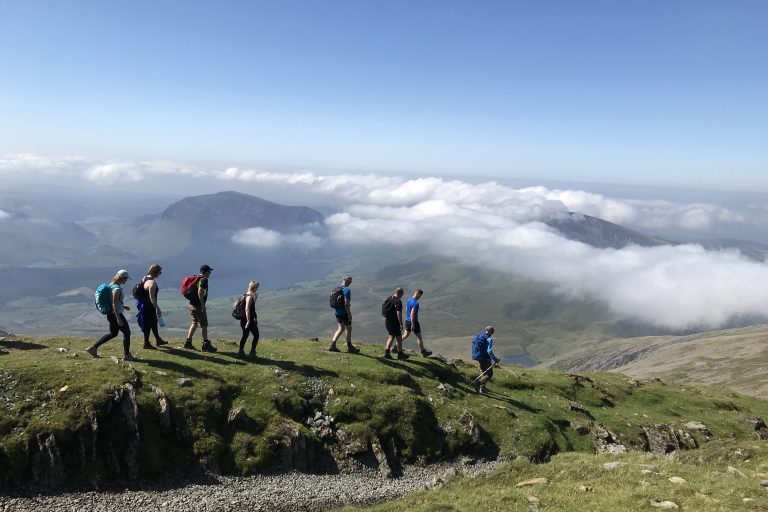 walkers with rucksacks decending a hill with peaks and valley in the distance